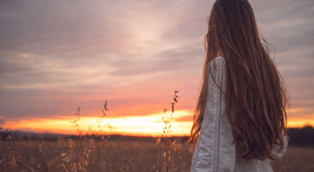 A woman with long hair stands in a field, silhouetted against a vibrant sunset sky.